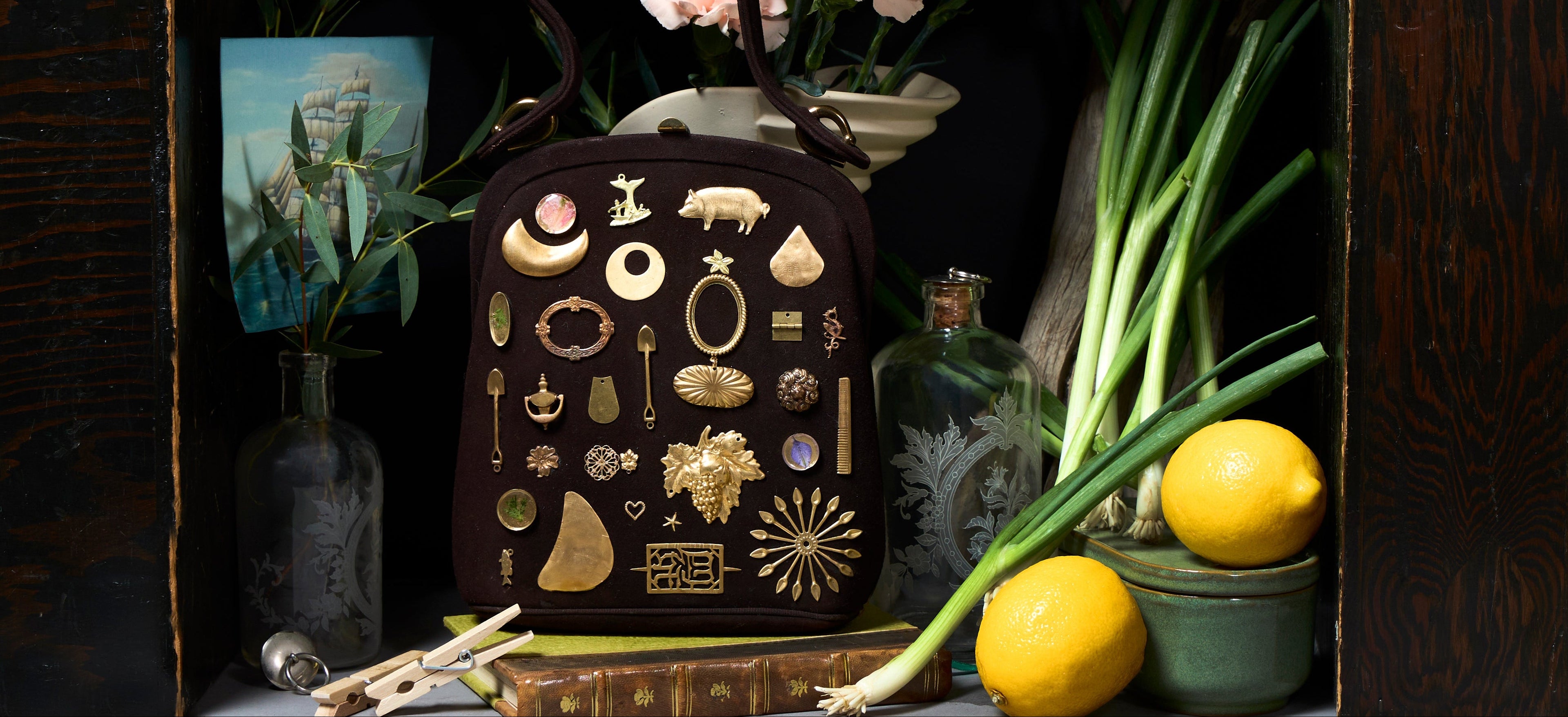 A dark brown handbag covered in gold-toned jewelry and accessory charms, styled in a rustic setting with lemons, green onions, flowers, and books.